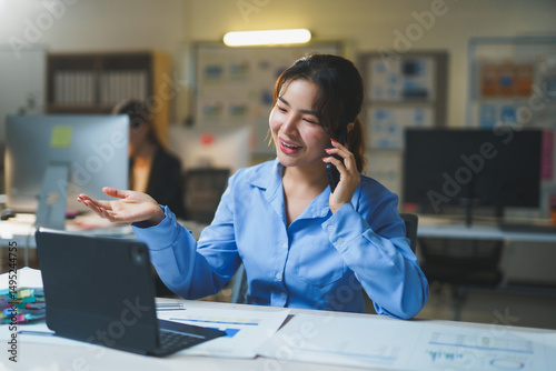 Young Asian businesswoman talking on mobile phone and working with digital tablet and financial documents in modern office at night