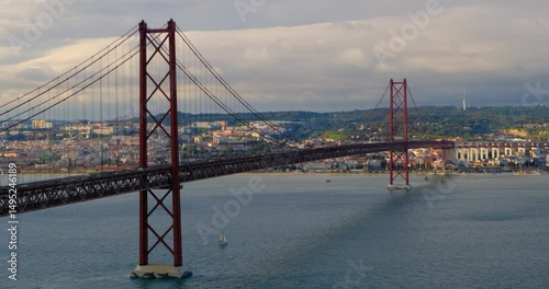 The iconic 25 de Abril Bridge stretches across the Tagus River in Lisbon, Portugal, its bold red structure cutting through soft afternoon light. Sailboats drift calmly, contrasting urban sprawl behind