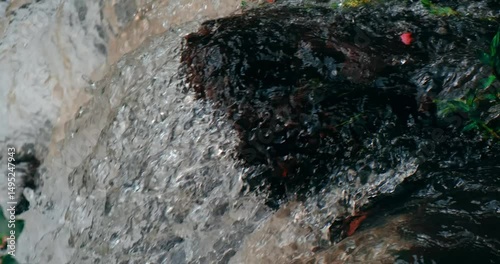 Crystal clear river water cascades over smooth rocks in a small, serene waterfall. Captured in slow motion, flowing water glistens under natural light, highlighting purity and tranquility. Close up.