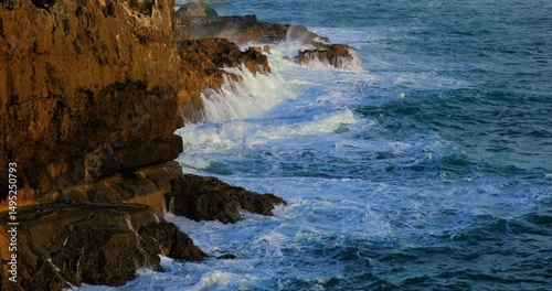 Powerful Atlantic Ocean waves crash against the rugged, sunlit cliffs of Cascais, Portugal. The golden hour light enhances the drama, showcasing nature’s force, sound, and coastline’s wild beauty.