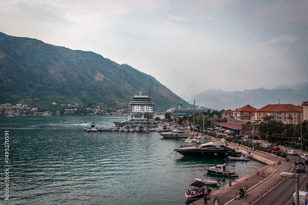 Naklejka premium Kotor, Montenegro. Town with a large cruise ship docked at a marina, surrounded by smaller boats, with mountains in the background and a bustling waterfront area