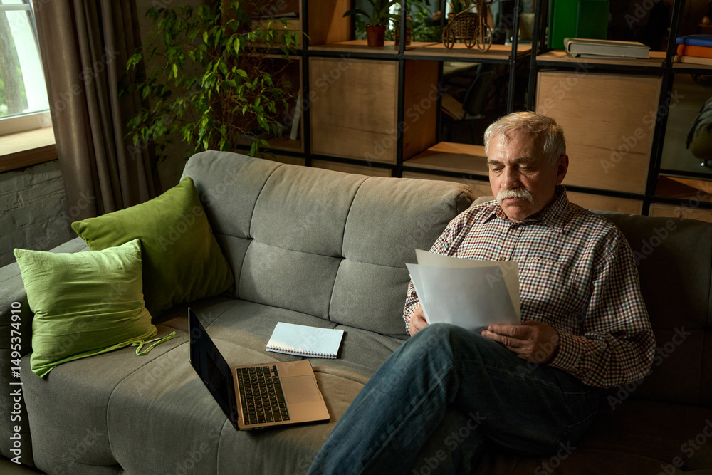 Fototapeta premium Senior man lying back on sofa and reading papers next to laptop and notebook in cozy living room. Comfortable education for seniors. Concept of elderly lifestyle, education, knowledge.