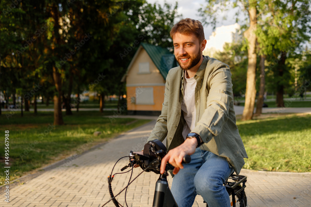 Obraz premium Young man enjoys a sunny day biking through a park with green trees and a residential building nearby