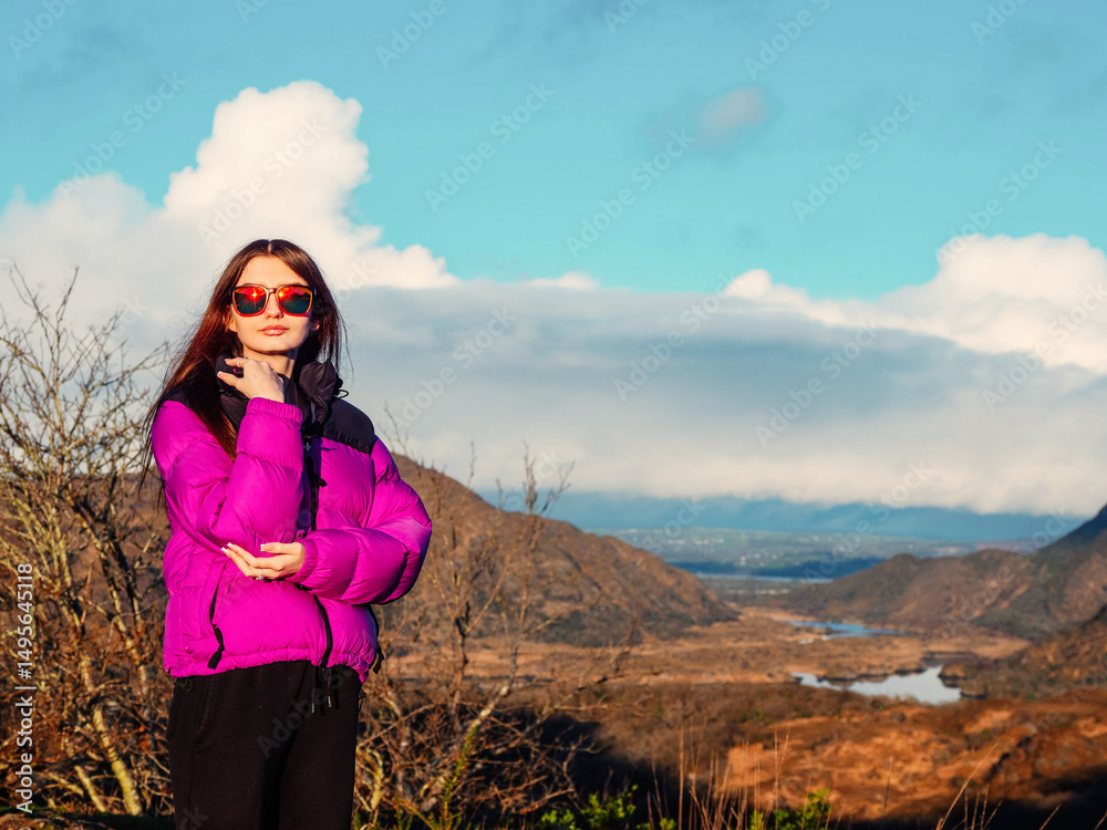 Naklejka premium A young teenager girl in a pink jacket stands on a rocky hillside. The sky is cloudy and the mountains in the background are covered in trees. Killarney area, Ireland. Travel and tourism.