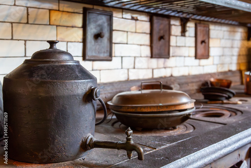 A large black Victorian kettle rests on an ancient iron range beside a copper pan in a traditional hearthside kitchen