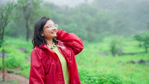 Portrait of beautiful Indian girl enjoying rain while standing in forest. Carefree female tourist in rain. Rain drops falling on face. Positive girl smiling and enjoying the rain outdoors.
