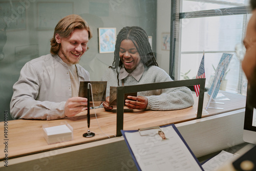 Smiling couple sitting at counter of public office, filling out forms and holding ID cards while engaging with official sitting behind glass partition