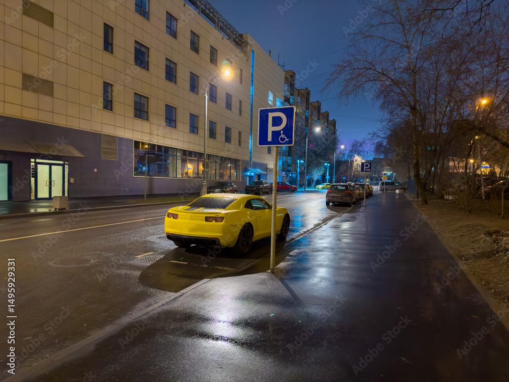 Fototapeta premium A vibrant yellow sport car is conspicuously parked on a wet city street at dusk, reflecting the glowing streetlights and the illuminated facade of a modern building. The low-angle shot captures the