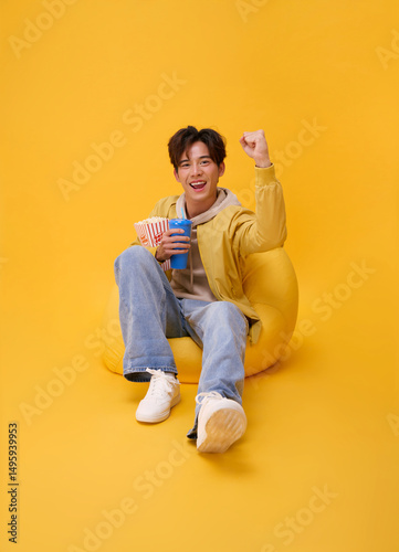 Happy young asian man sitting on bean bag eating popcorn and watching movie at the cinema home studio isolated on yellow background.
