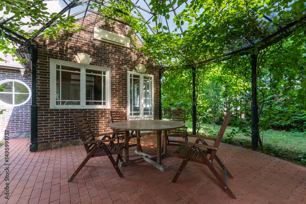 Obraz premium A beautiful ivy-covered gazebo with garden furniture, in the backyard of a brick house on a sunny summer day.