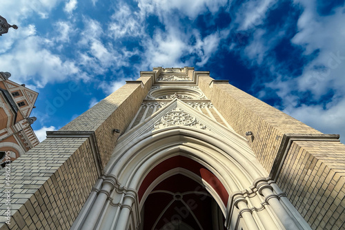 Upward view of the neo-Gothic The Name of Mary Church, or Novi Sad catholic cathedral , showing ornate brick tower, pointed arch portal and blue sky backdrop, a Vojvodina architectural heritage.