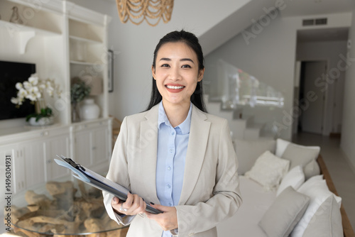 Portrait of smiling Vietnamese realtor woman holds folder, look at camera stands in modern living room. Professional real estate agent showcasing new property, selling luxury property, bank mortgage