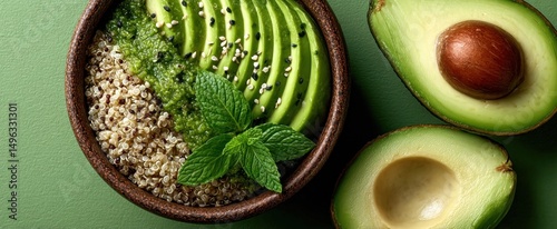A healthy bowl with sliced avocado, quinoa, mint leaves, and chia seeds beside a halved avocado on a green surface.