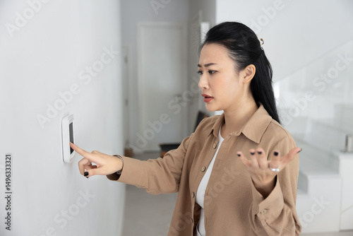 Woman standing indoors with concerned and frustrated expression while points at wall-mounted digital thermostat. Frozen or unresponsive screen, too hot or too cold, device malfunction, error message