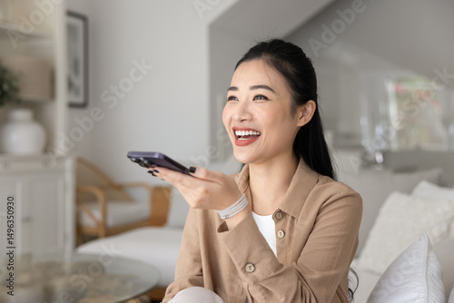 Young 30s Asian woman make hands-free phone call, asking virtual assistant for information using speakerphone seated on sofa in living room. Innovative tech, artificial intelligence, chat bot usage
