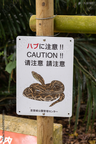 A caution sign at Shuri Castle warns visitors about the presence of habu snakes, featuring multilingual text and an illustration.