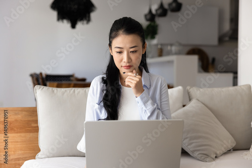 Photography Pensive Asian woman sits on sofa with laptop, solving work-related problem or challenge, search creative idea or solution, making decision, thinks on task to complete at home