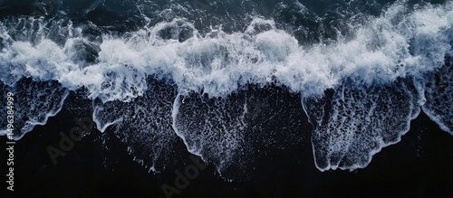 Drone aerial view of the sea or ocean waves splashing on a dark navy blue or gray sand beach with beautiful transparent clear water