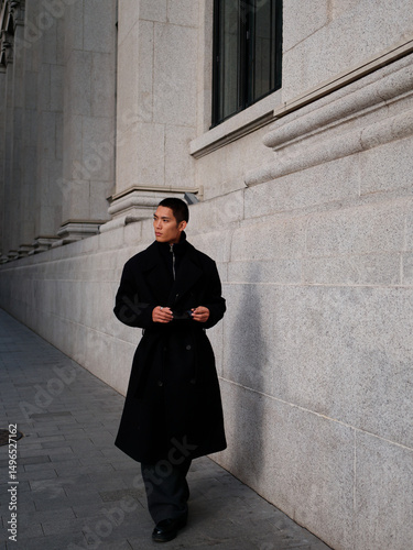 Portrait of handsome Chinese young man wearing black overcoat posing in the street, young guy with black short hair all in black with urban background. Male fashion, cool Asian young man lifestyle.