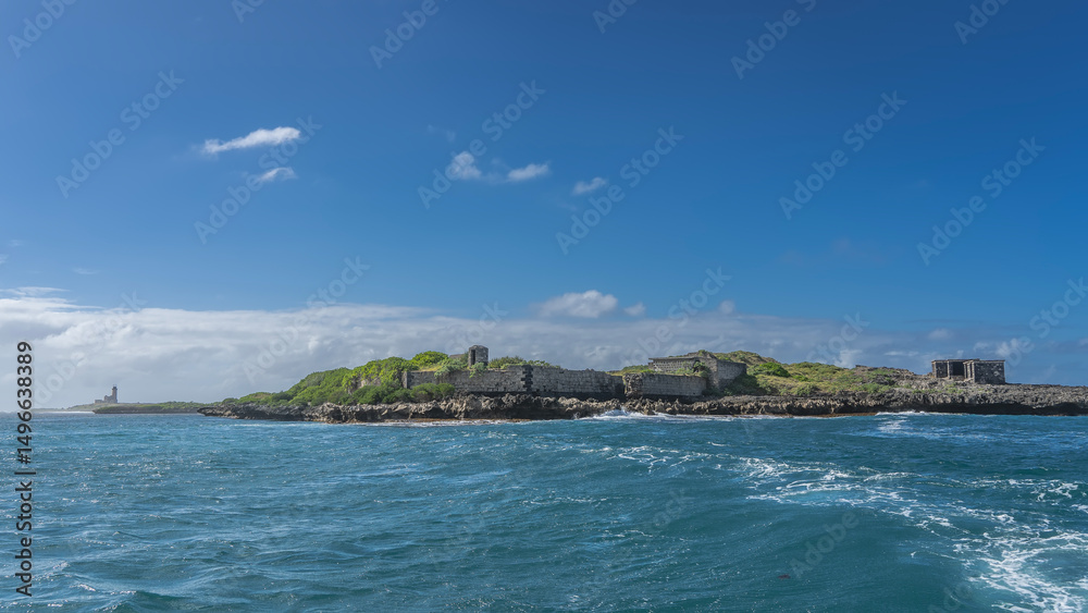 Obraz premium A small island in the ocean. The green hills and ruins of old buildings. Aquamarine water, blue sky with white clouds. Mauritius. Prison Island