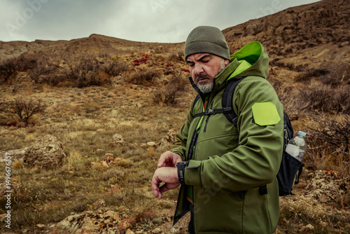 Fotografie Man checks watch while hiking in rugged terrain during overcast weather in a mou
