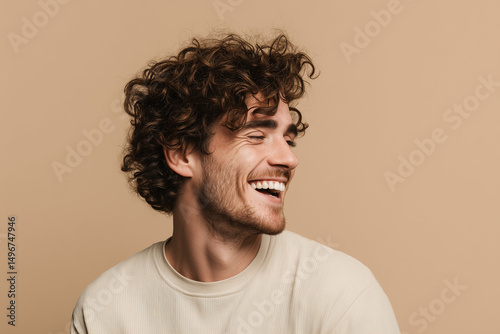 Portrait of a cheerful young man with curly hair laughing with closed eyes isolated over beige background