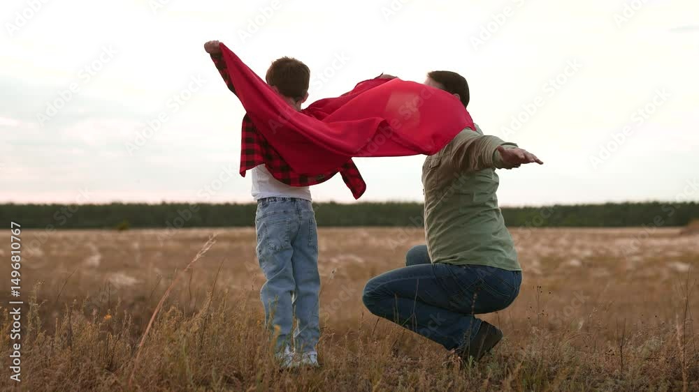 Cheerful father and son play superhero with red cape simulating flight in countryside in field. Father and son with red cape feel lightness and freedom. Support from father to son with red cape