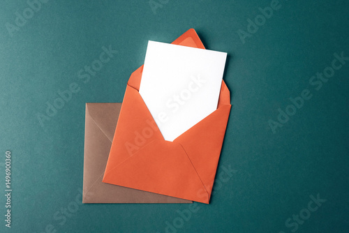 Orange and brown envelopes with blank white card on dark green background. Top view, flat lay, mockup
