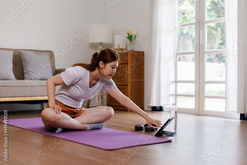Young Asian woman stretching yoga workout on exercise mat while online training class with computer laptop at home in living room
