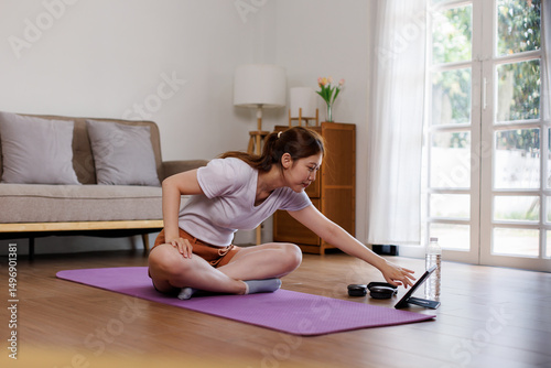 Young Asian woman stretching yoga workout on exercise mat while online training class with computer laptop at home in living room
