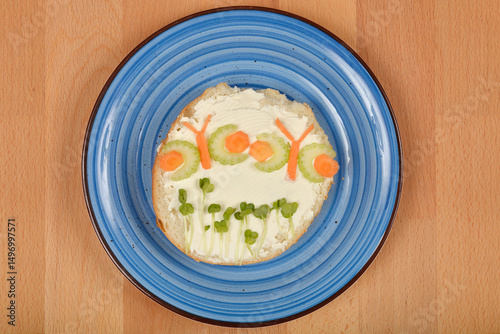A creatively decorated open-faced sandwich, featuring cream cheese, sprout "grass," and vegetable "butterflies" made from celery and carrot, rests on a blue plate atop a wooden surface