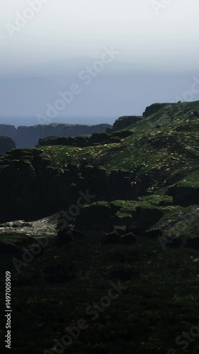 Misty conditions create an atmospheric view of rocky cliffs and lush green hills along a rugged coastline at dusk. The soft light enhances the serene and mysterious landscape. photo