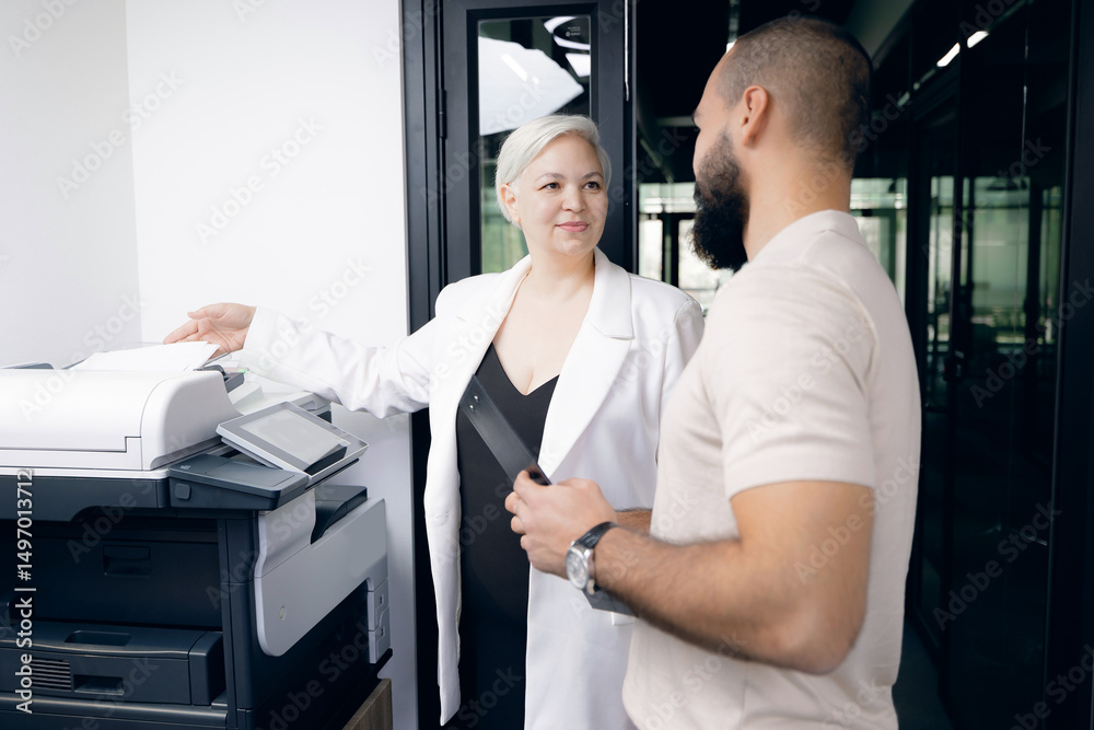 Obraz premium Two colleagues engaged in conversation beside printer in modern office. one woman in white coat with short grey hair, and man with beard in casual attire.