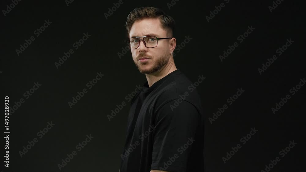 Young man with beard and glasses stands confidently against a black wall with an isolated and focused demeanor. Young man with beard and glasses stands confidently against a black wall with an isolated and focused demeanor.