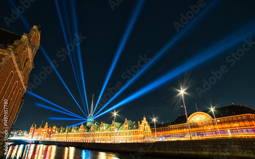 The Børsen or Borsen building in Copenhagen glows at night with blue light beams radiating from its iconic spire, blending historic architecture and modern light art along a vibrant, reflective canal photo