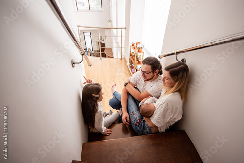 Young family with newborn baby and daughter are sitting on stairs in their modern home, talking and pointing at something upstairs, enjoying their time together