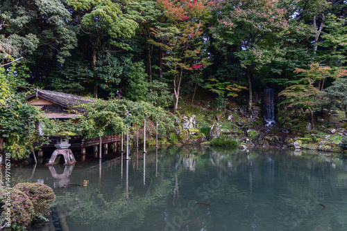 Traditional pavilion surrounded by greenery and autumn foliage beside a quiet pond with reflections and a waterfall in Kenrokuen Garden, Kanazawa, Japan