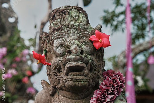 Balinese stone statue adorned with colorful flowers in a lush garden setting during sunset in Bali, Indonesia