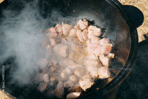 Pieces of meat frying in oil in a large cast iron cauldron with steam. Cooking outdoor in a big pot for holiday.
