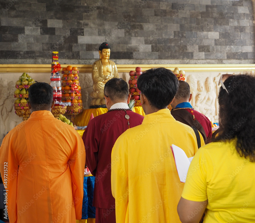 Fototapeta premium A group of individuals in yellow and orange robes stand facing a shrine featuring a Buddha statue. Offerings of fruit are stacked around the statue. The setting suggests a Buddhist temple with a stone