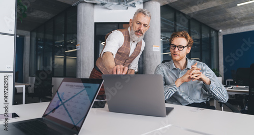 Konstfotografi Senior businessman guides young colleague by pointing at laptop screen during a