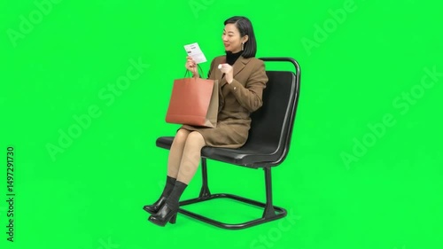 Elegant shopper enjoys their purchases on a modern chair in front of a vibrant green backdrop, smiling brightly. 