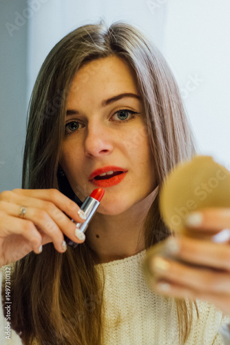 Woman applying red lipstick while looking in a compact mirror, beauty routine and cosmetic product concept.