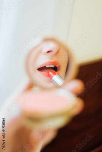 Woman applying vivid red lipstick using a compact mirror. Reflection of a female during morning makeup routine. Beauty procedure for cosmetic product.