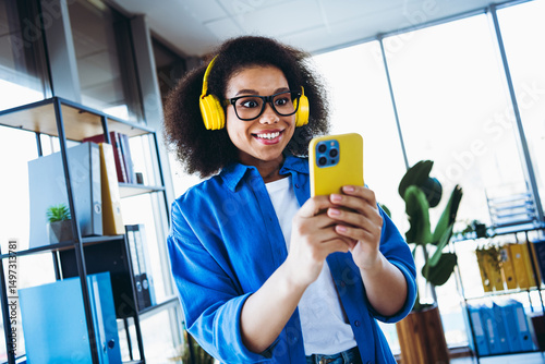 Young professional using mobile in modern office space wearing casual attire and headphones while expressing joy photo