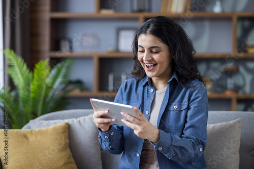A smiling woman enjoys time on her tablet, relaxing on a comfortable sofa at home with a plant.