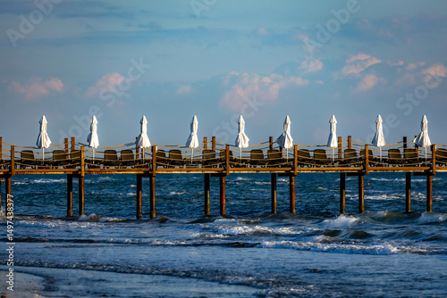 Wooden beach pier with rows of closed white umbrellas and sunbeds in warm evening light before tourist season. Antalya, Mediterranean coast, Turkey