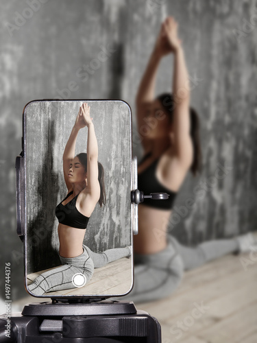 Fitness influencer doing home workout and stretching on yoga mat while live streaming on smartphone. Sporty young woman filming online fitness training for social media followers.