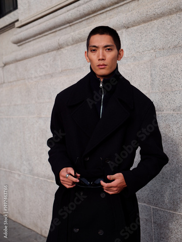 Portrait of handsome Chinese young man wearing black overcoat posing in the street, young guy with black short hair all in black with urban background. Male fashion, cool Asian young man lifestyle.