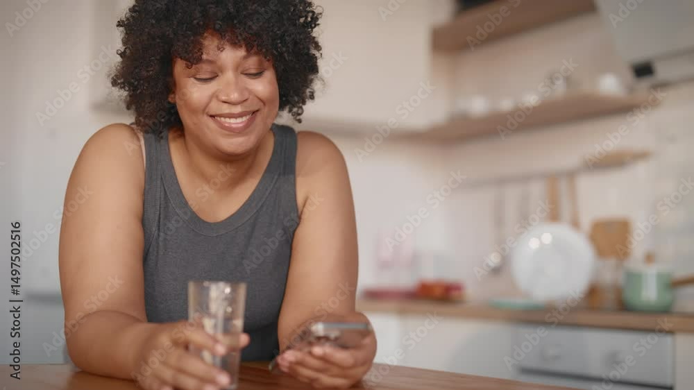 Pretty young black woman reading funny news and watching videos in smartphone. Charming african american lady drinking water in home kitchen in morning, useful habits for health, healthy lifestyle